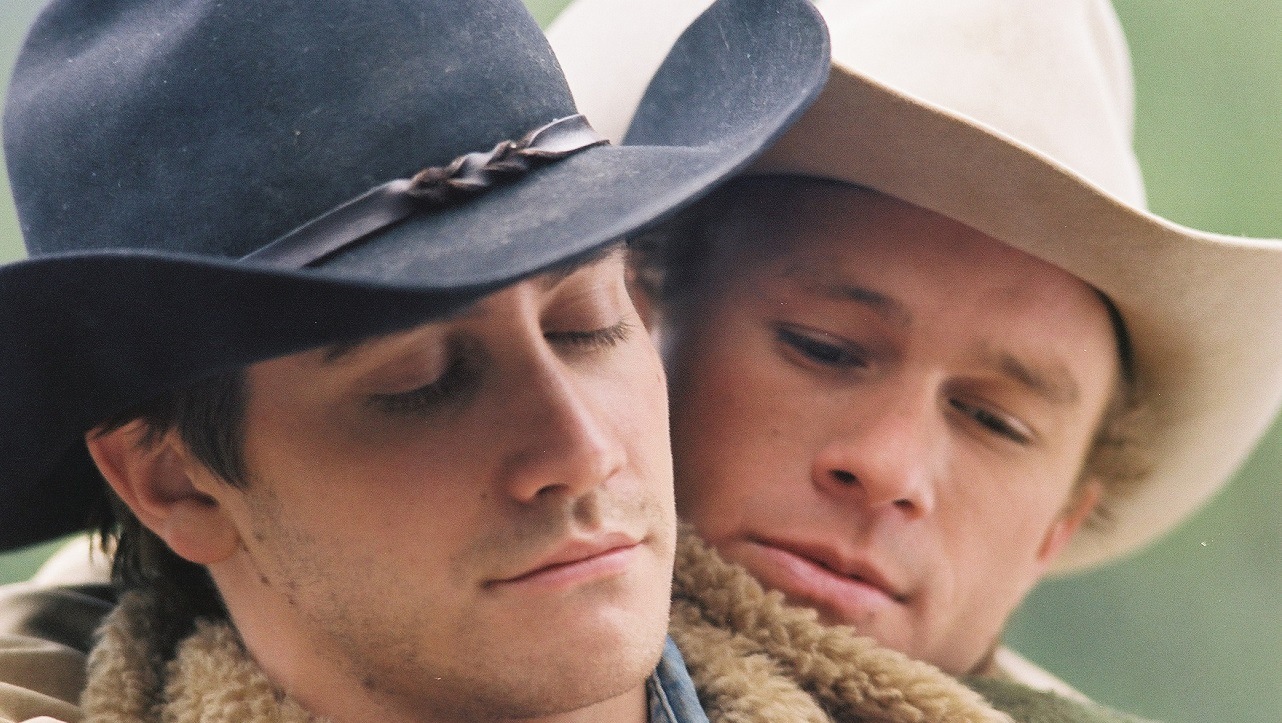 dois homens jovens brancos de chapéu de caubói numa momento afetuoso: um abraça o outro por trás. Seus rostos estão em close.